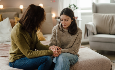 A mother holds her teen daughter's hand while talking on a bed